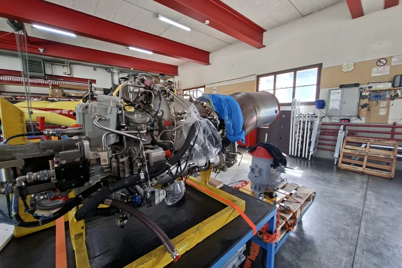 Hubschraubertriebwerk auf einem Montagegestell in einer hellen Werkstatt, mit einem Hubschrauber im Hintergrund.