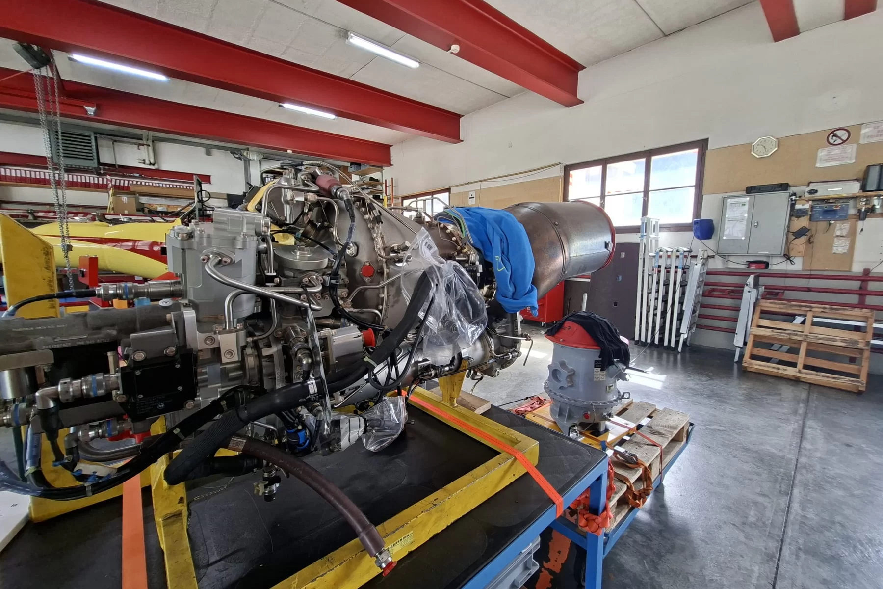 Hubschraubertriebwerk auf einem Montagegestell in einer hellen Werkstatt, mit einem Hubschrauber im Hintergrund.